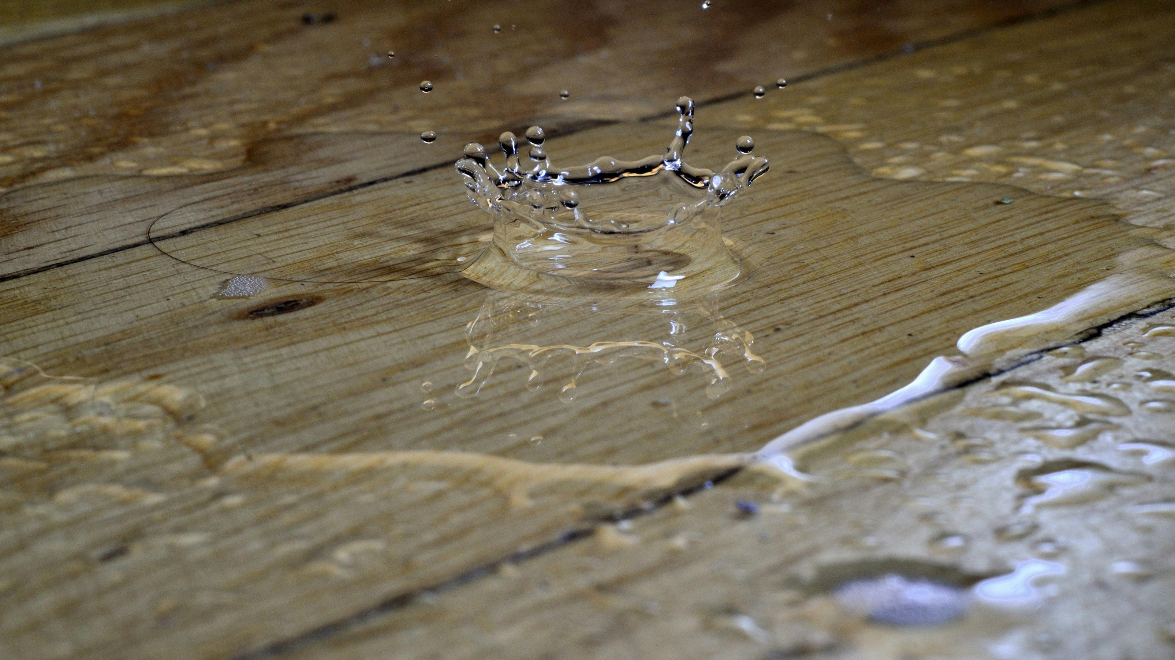 Dynamischer Wassertropfen-Aufprall auf einer Holzoberfläche mit charakteristischen Spritzern und Wasserkrone, die sich in kristallklaren Tropfen gegen die natürliche Holzmaserung abzeichnet.