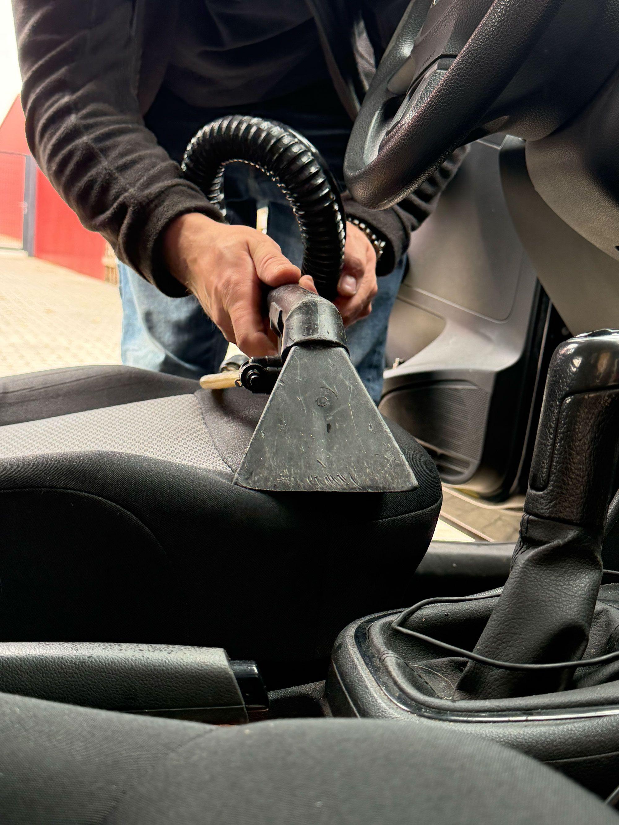 Person reinigt grauen Autositz mit Polsterreinigungsgerät, dreieckiger Saugaufsatz im Einsatz, Lenkrad und Schaltknauf im Hintergrund.