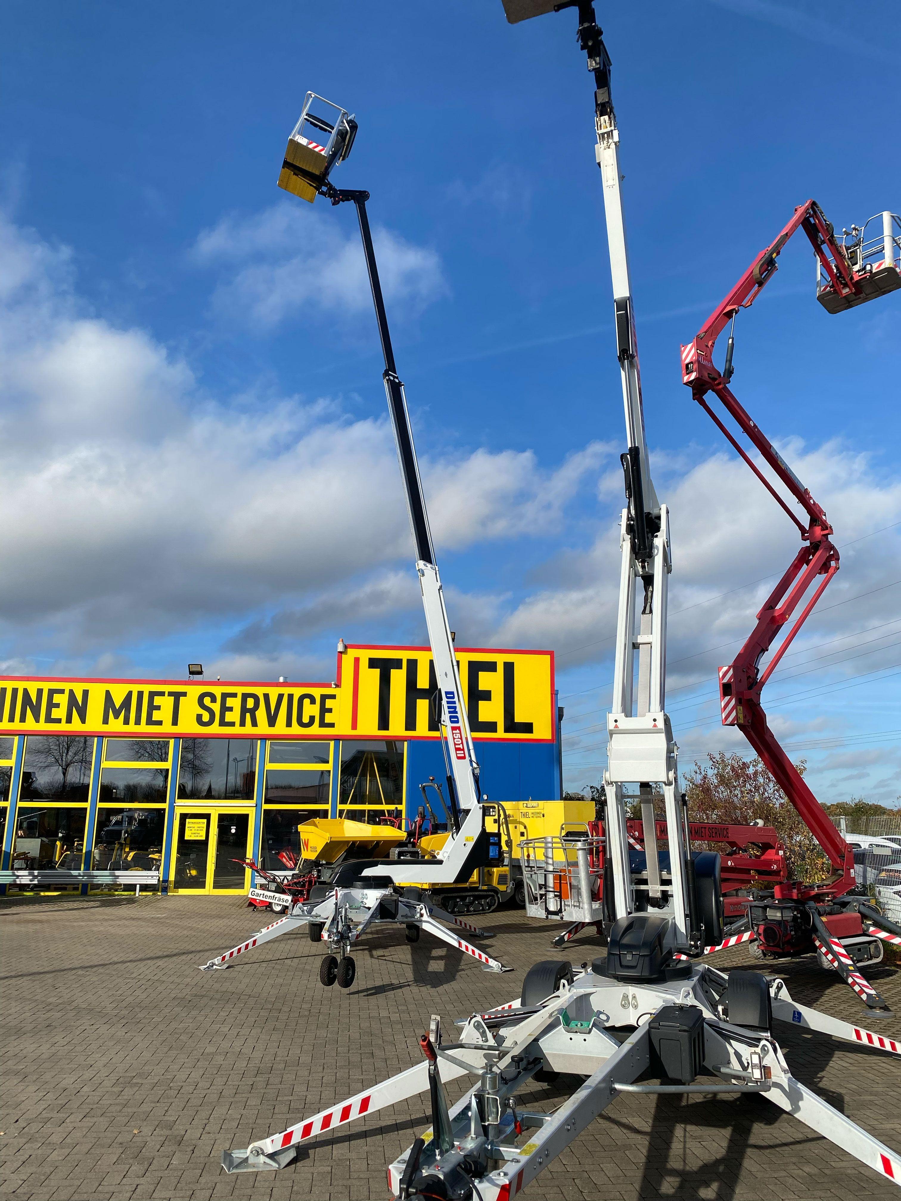 Verschiedene Arbeitsbühnen unterschiedlicher Bauart und Höhe auf einem Ausstellungsgelände vor gelbem Thiel-Firmengebäude, darunter weiße Anhänger-Arbeitsbühnen im Vordergrund und rote sowie weiße Teleskop- und Gelenkteleskopbühnen.